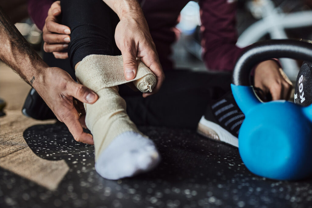 Close up of unrecognizable coach bandaging injured athlete's ankle in a gym.